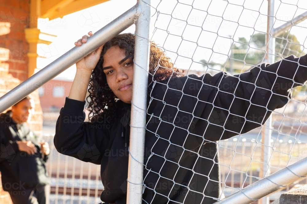 teen girl wearing black looking out past chainlink fence with another person in background - Australian Stock Image