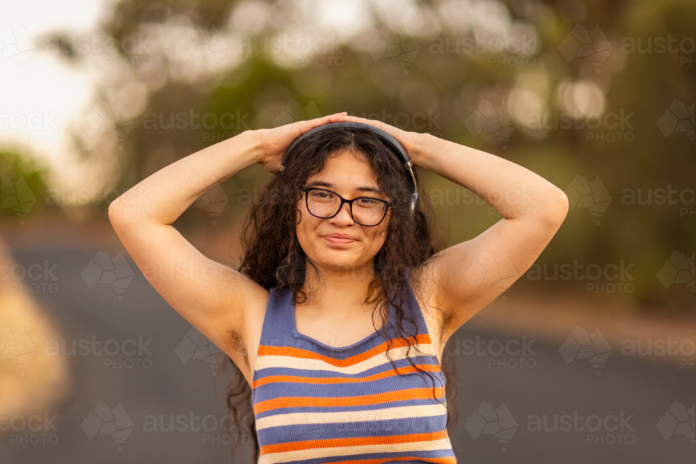teen girl wearing headphones and glasses with hands on her head showing hairy armpits - Australian Stock Image