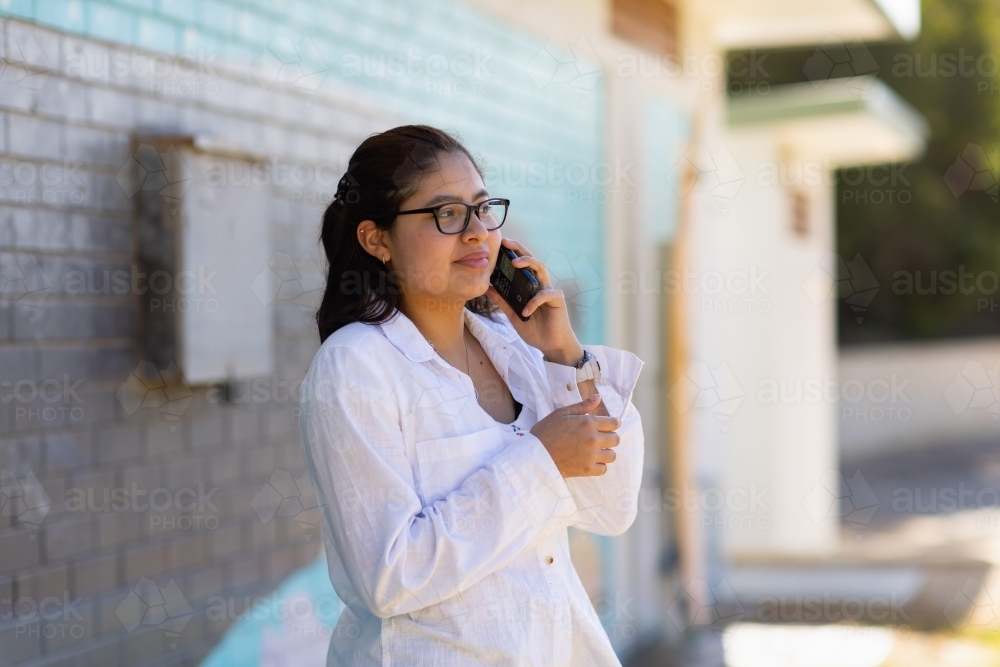 teen girl wearing white shirt talking on phone standing near painted brick wall - Australian Stock Image