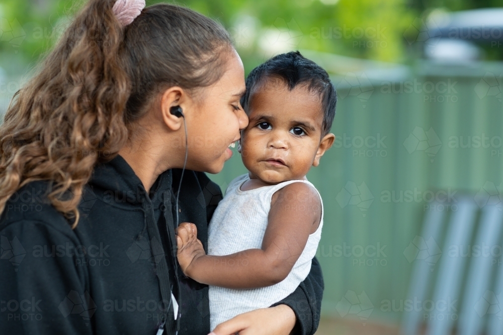 Teen girl with earbuds in ears holding and cuddling baby - Australian Stock Image
