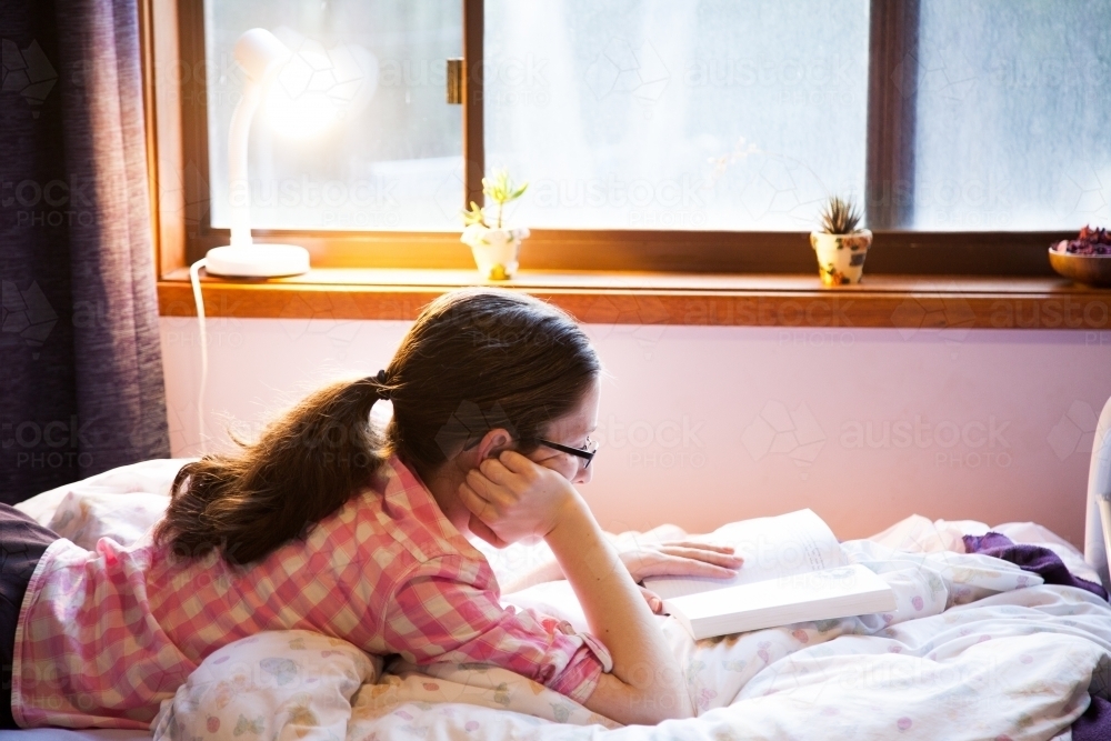 Teen person lying on her bed reading a fiction book with lamp on - Australian Stock Image