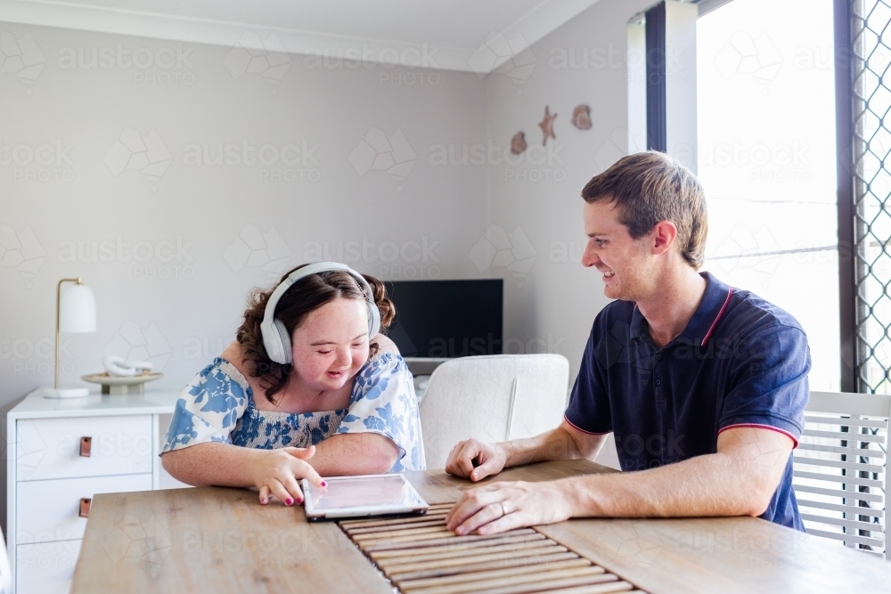 Teen wearing headphones talking with adult disability support worker in home office using tablet - Australian Stock Image