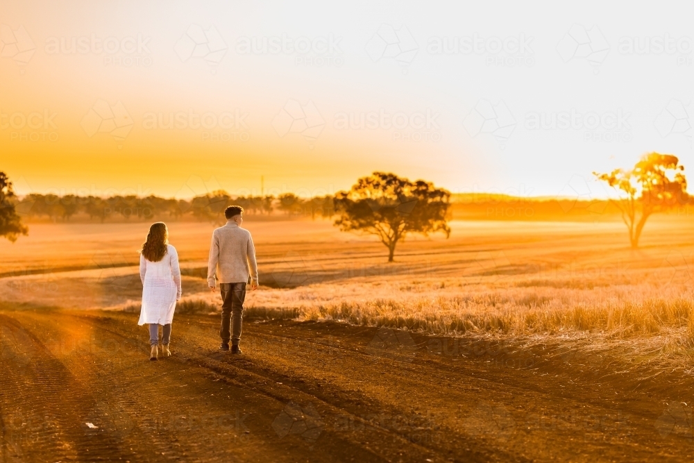 Teenage boy and girl walking together on rural dirt road at sunset - Australian Stock Image