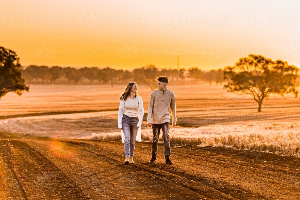Teenage boy and girl walking together on rural dirt road at sunset - Australian Stock Image
