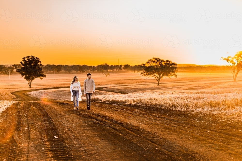 Teenage boy and girl walking together on rural dirt road at sunset - Australian Stock Image