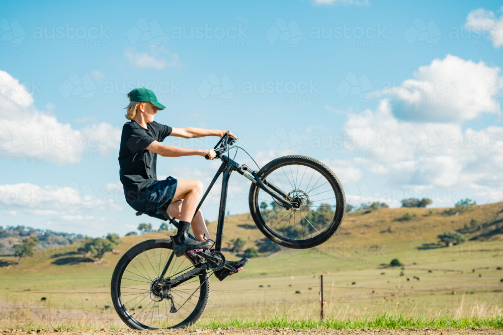 Teenage boy doing wheelie on mountain bike on remote country road - Australian Stock Image