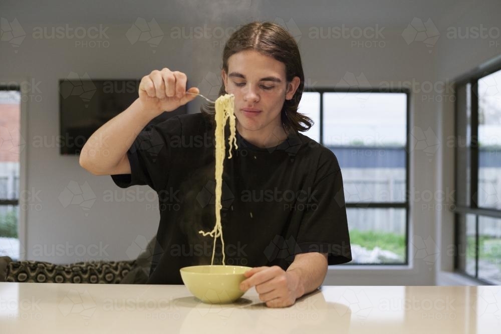Teenage boy eating noodles at a kitchen bench - Australian Stock Image