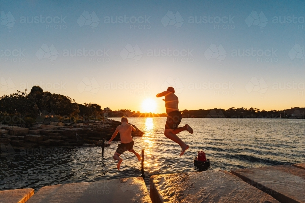 Teenage boy jumping from ledge into water in bright setting sun - Australian Stock Image