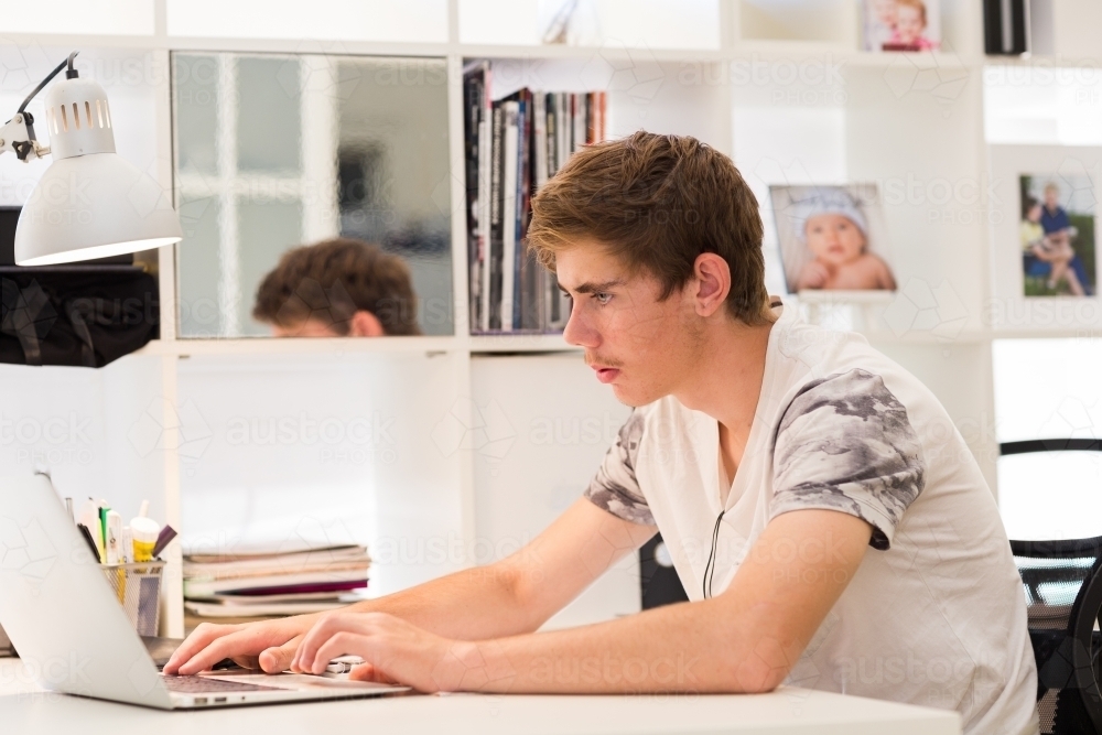 Teenage boy sitting at desk typing on laptop computer - Australian Stock Image