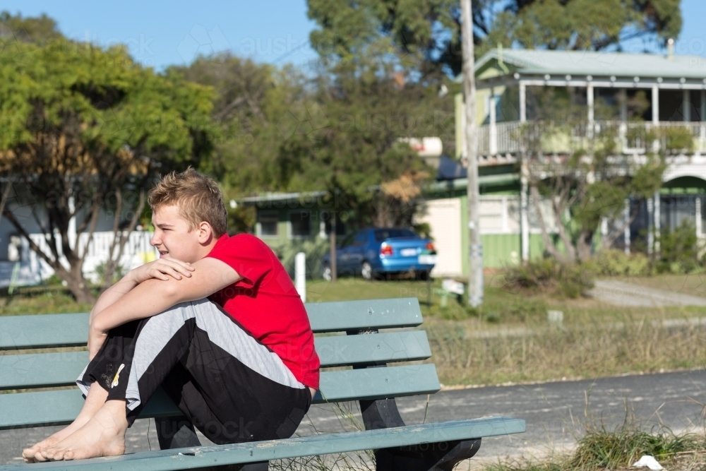 Teenage boy sitting on park bench with arms around knees - Australian Stock Image