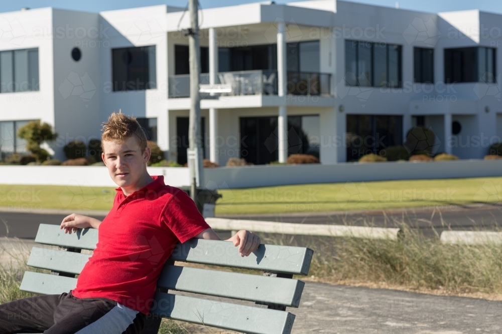 Teenage boy sitting on park bench, with mansion in the background - Australian Stock Image