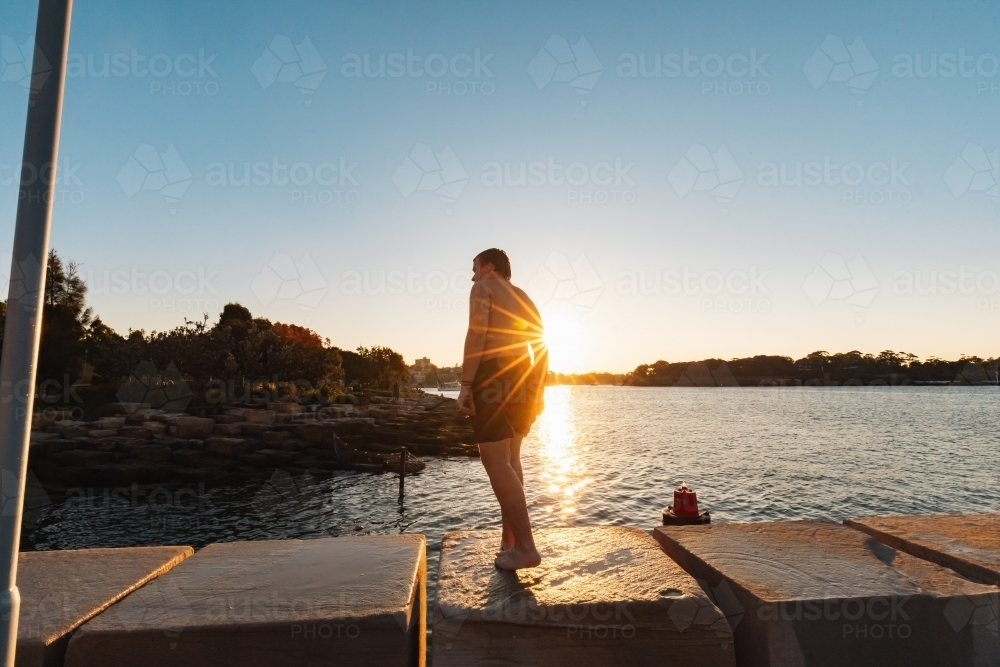 Teenage boy standing on ledge by water in bright setting sun - Australian Stock Image