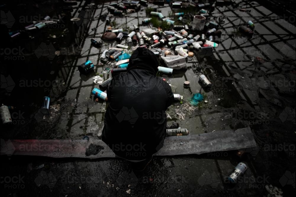 Teenage boy surrounded by empty spray cans in deserted warehouse - Australian Stock Image