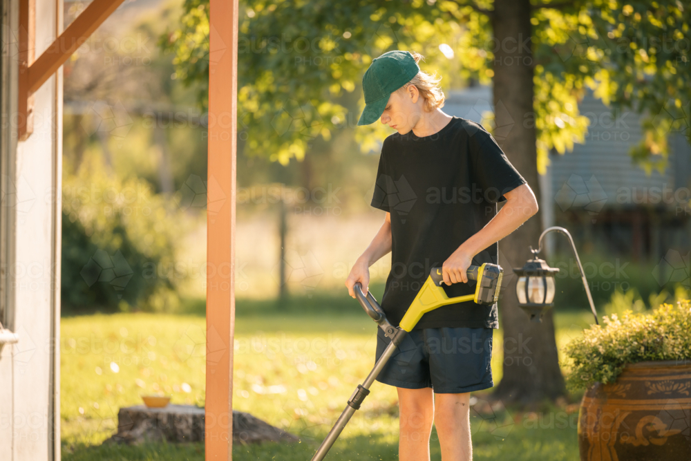 Teenage boy using line trimmer on lawn edging in country garden - Australian Stock Image