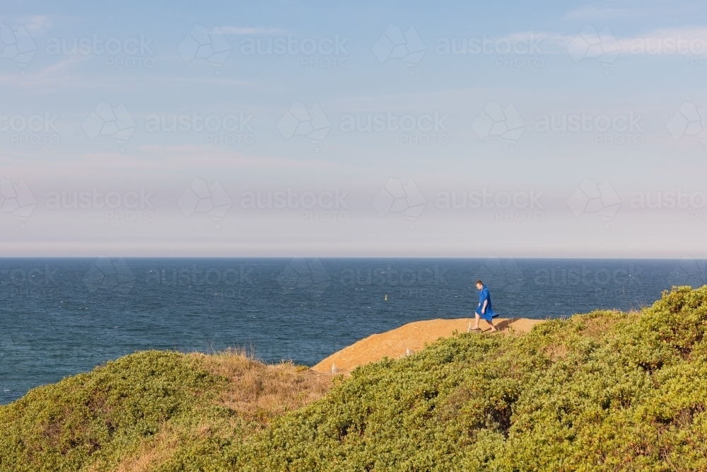 Teenage boy wearing hooded towel walking along cliff edge with ocean background - Australian Stock Image