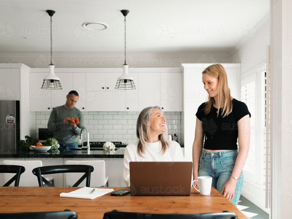 Teenage daughter giving mum a cup of coffee working on her laptop on wooden table - Australian Stock Image