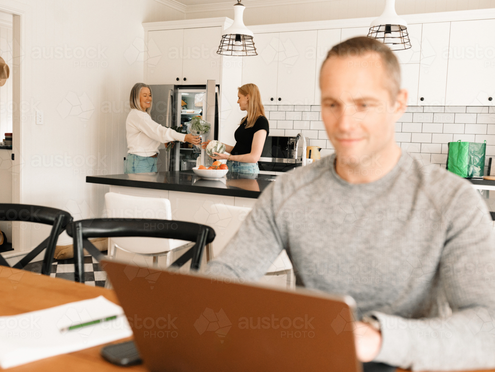 Teenage daughter helping mum with groceries in the kitchen while dad works on computer - Australian Stock Image