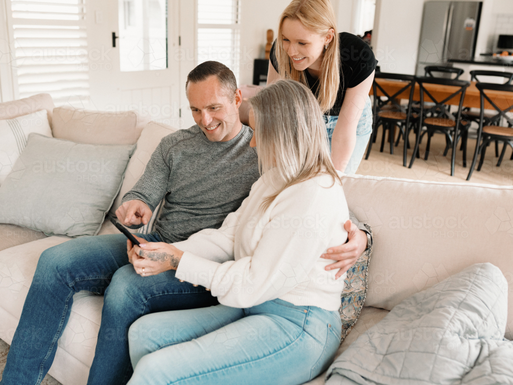 Teenage daughter leaning over her parents seated on the lounge looking at fold phone screen - Australian Stock Image