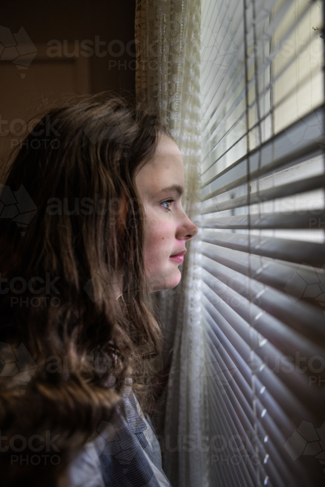 Teenage girl at window looking out venetian blinds - Australian Stock Image