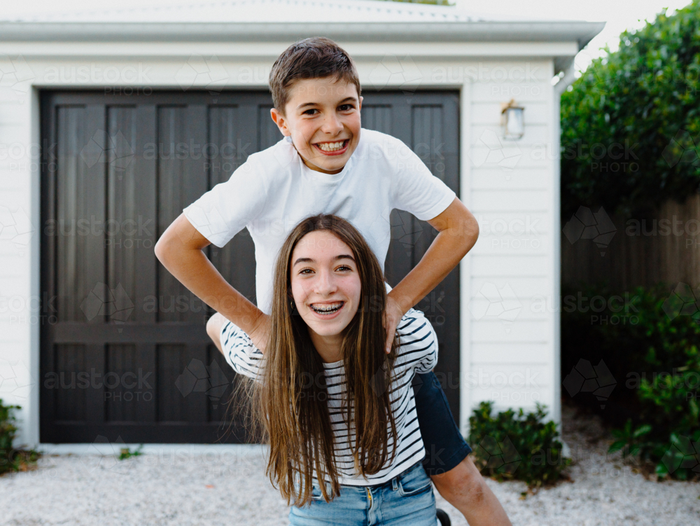 Teenage girl doing piggyback with brother on her back in their yard. - Australian Stock Image