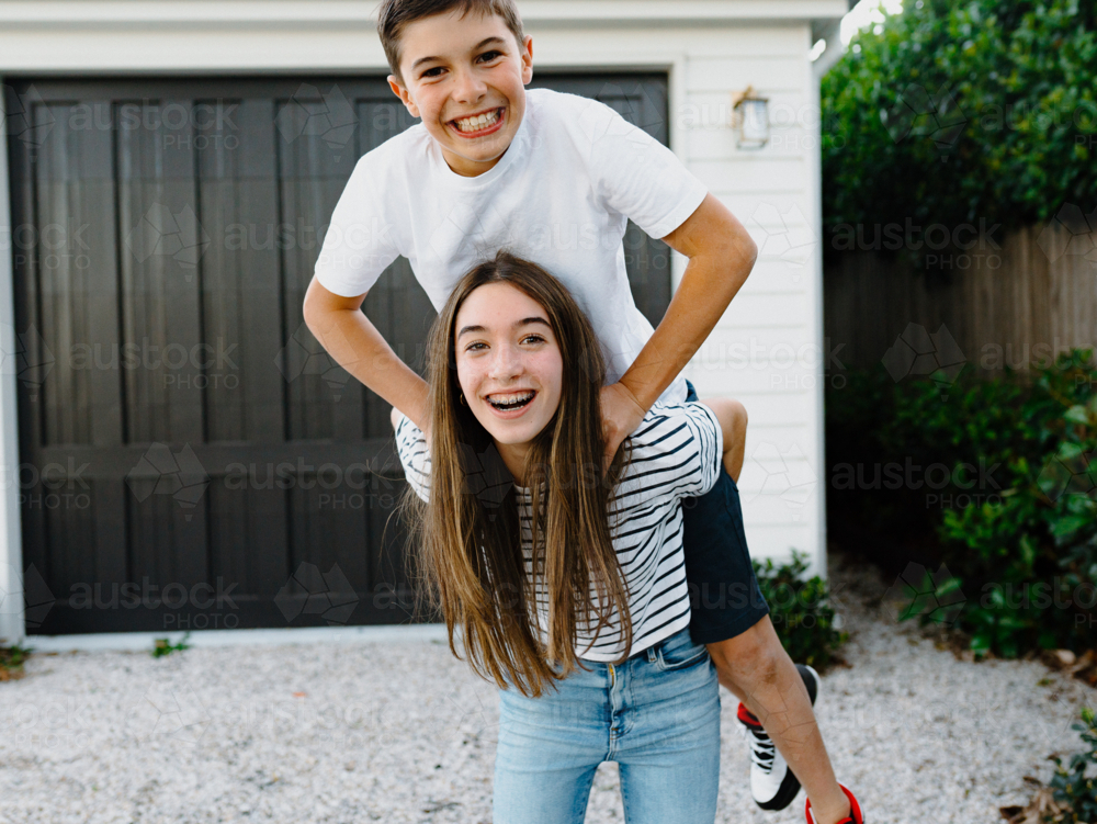 Teenage girl giving her brother a piggyback around their yard at home - Australian Stock Image