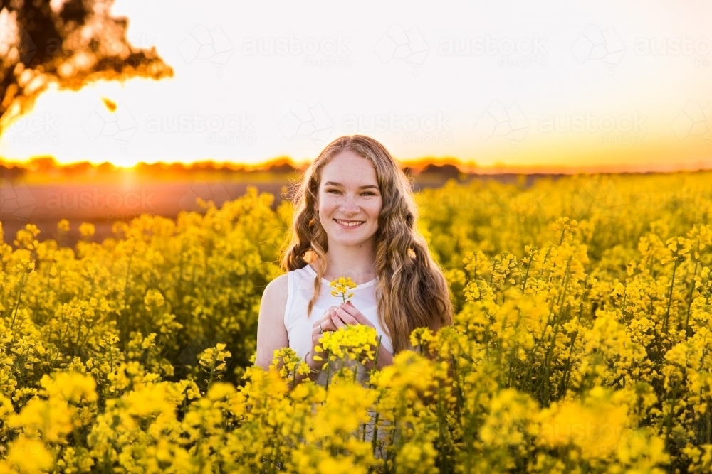 Teenage girl in field of canola on farm at sunset smiling - Australian Stock Image