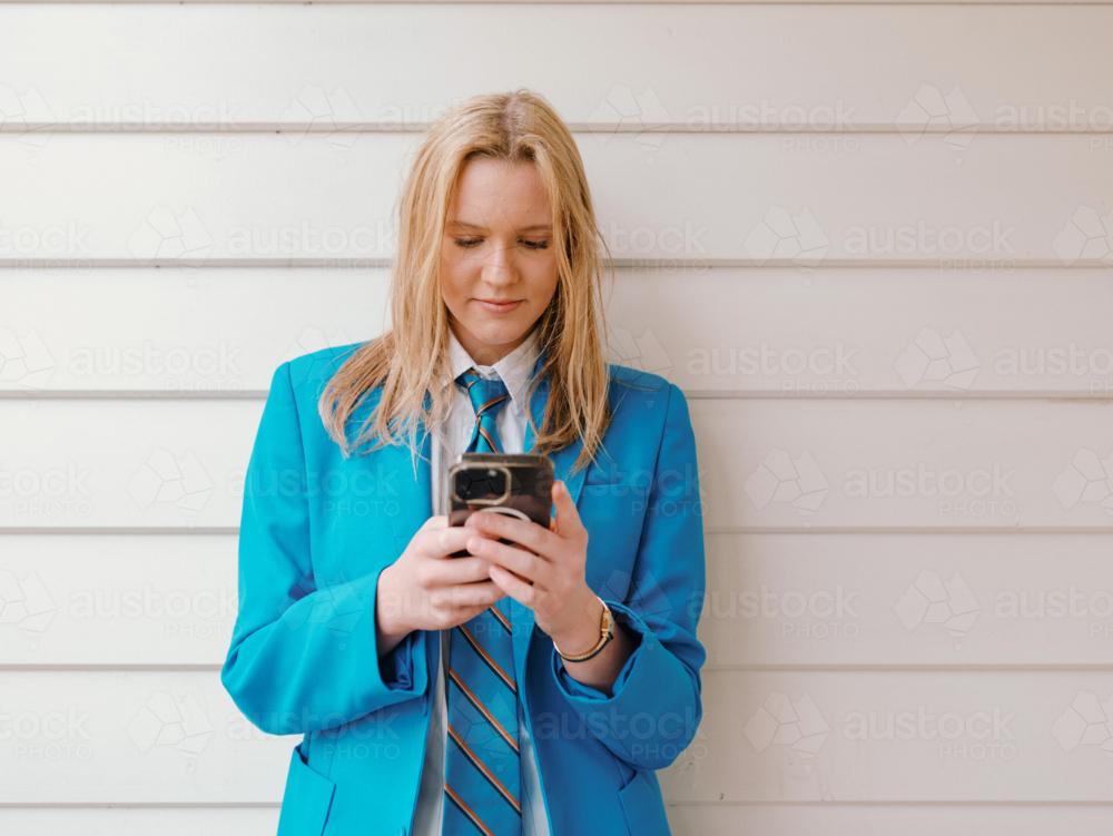 Teenage girl in school uniform leaning against the white wall while engaged on her phone - Australian Stock Image