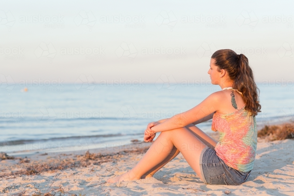 Teenage girl sitting on beach looking out to sea - Australian Stock Image