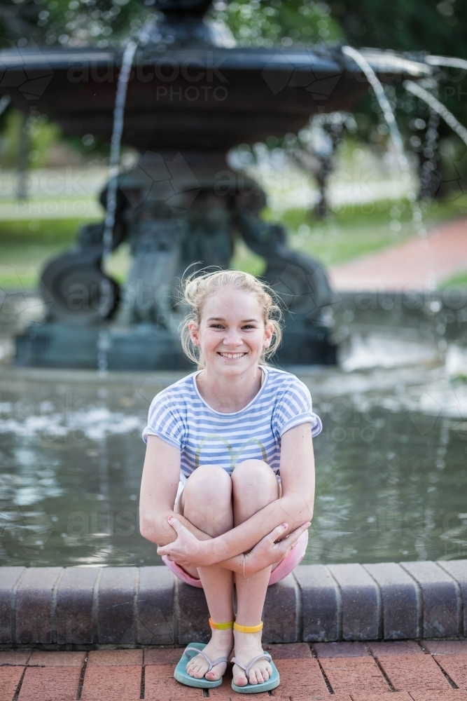 Teenage girl sitting on edge of water fountain smiling - Australian Stock Image