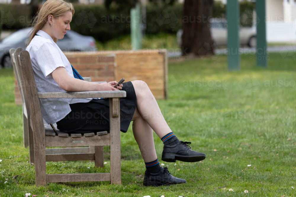 Image of Teenage girl sitting on wooden bench outdoors scrolling on her ...