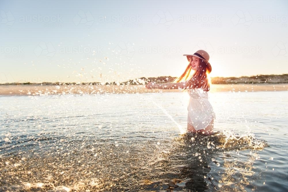 Teenage girl splashing water at the beach with the setting sun behind her - Australian Stock Image