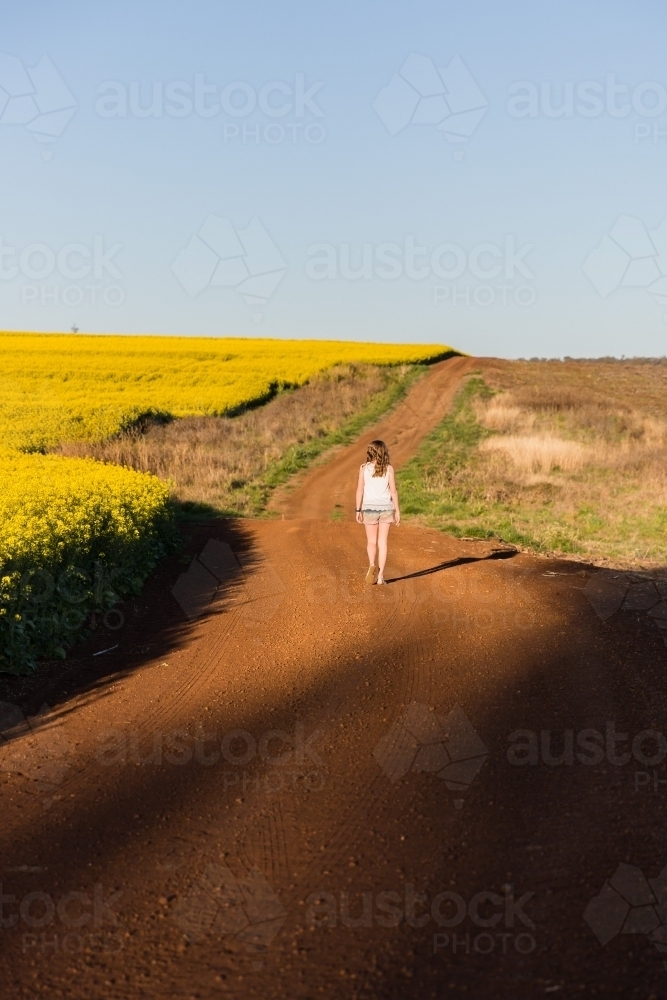 Teenage girl walking down dirt road on farm with canola crop - Australian Stock Image