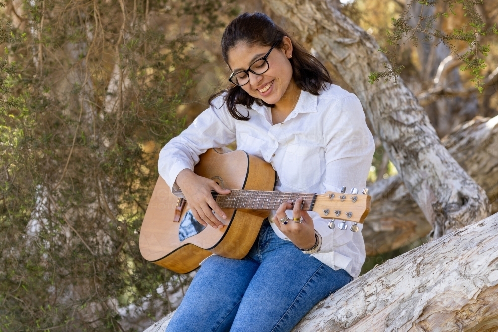 teenage girl wearing glasses outdoors playing acoustic guitar in tree - Australian Stock Image