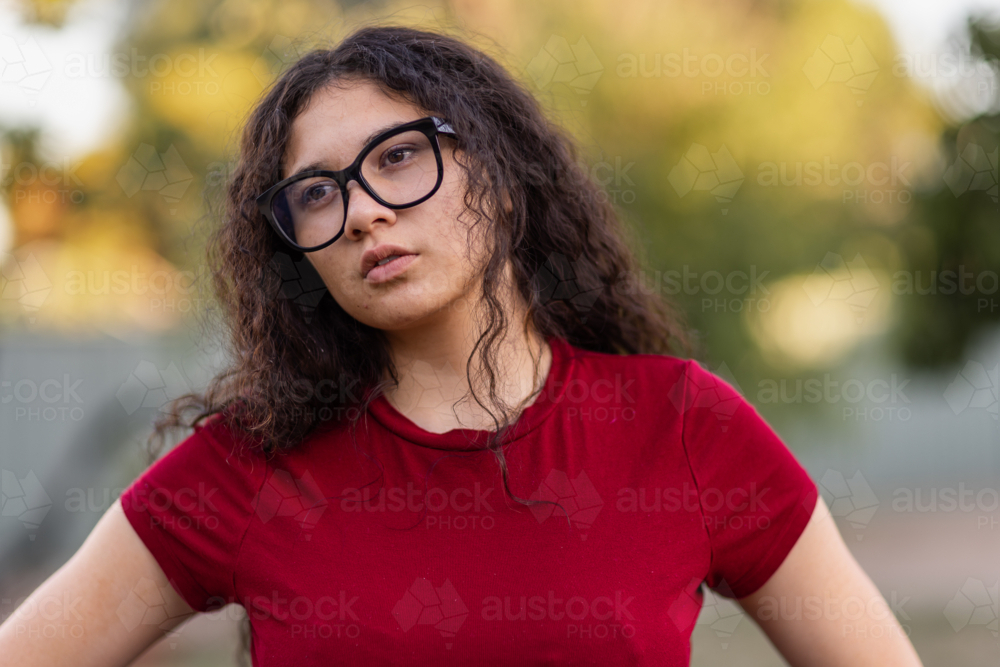 Teenage girl with dark curly hair wearing glasses and red shirt standing outdoors - Australian Stock Image