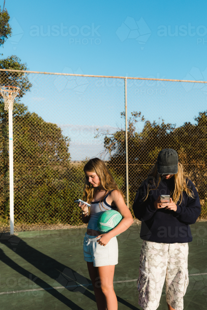 Teenage girls on smart phones while holding netball at outdoor court - Australian Stock Image