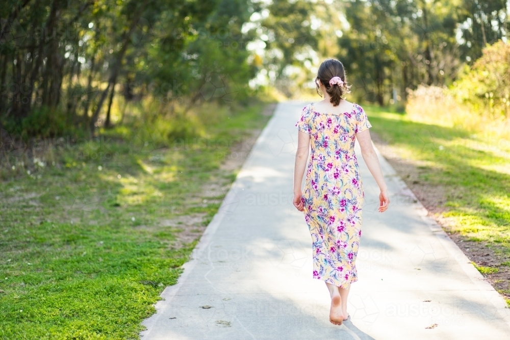 Teenage woman in floral dress in early Spring walking along a footpath through park in Singleton - Australian Stock Image