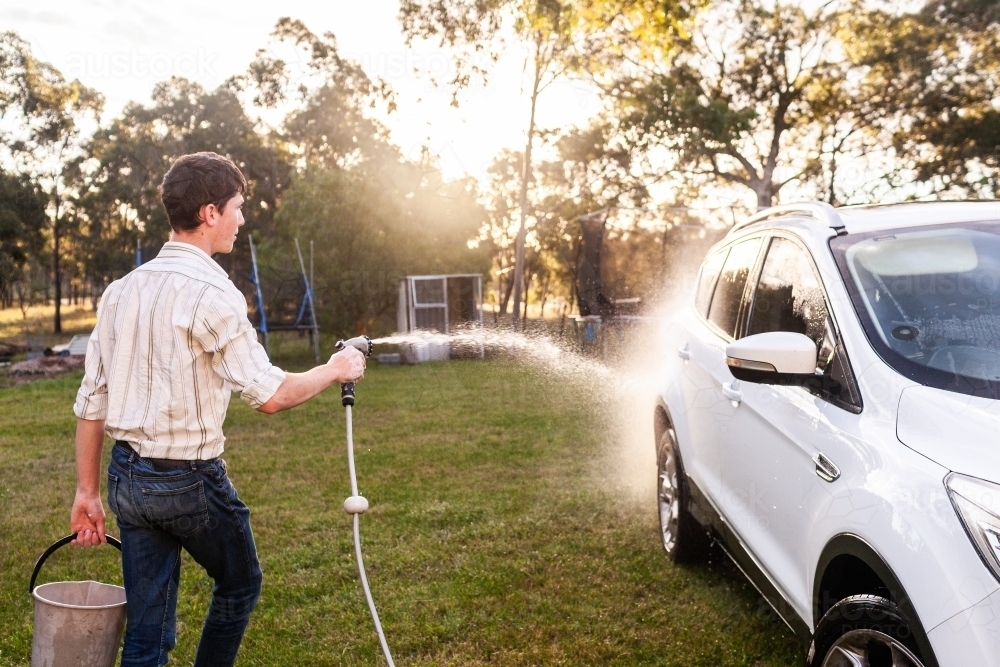 Teenager hosing suds of family car in sunset light - Australian Stock Image