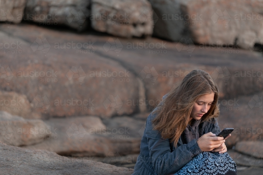 Teenager sitting outdoors at twilight and texting on smartphone - Australian Stock Image