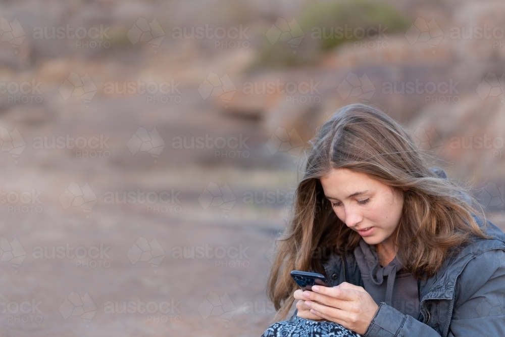 Teenager sitting outdoors at twilight and texting on smartphone - Australian Stock Image