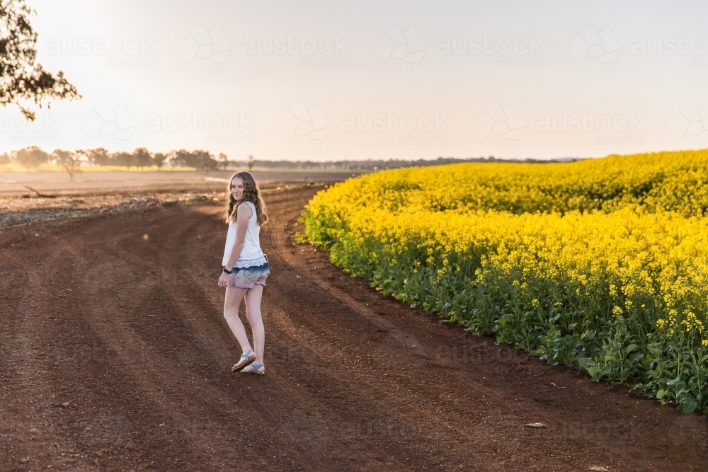 Teenager walking on dirt road on farm looking over shoulder next to paddock of canola - Australian Stock Image