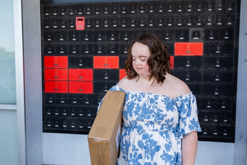 Teenager with down syndrome holding brown box parcel by post office looking down - Australian Stock Image