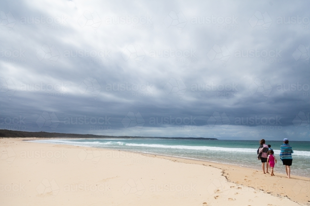 Teenagers and children walking towards the sea on beach under stormy clouds - Australian Stock Image