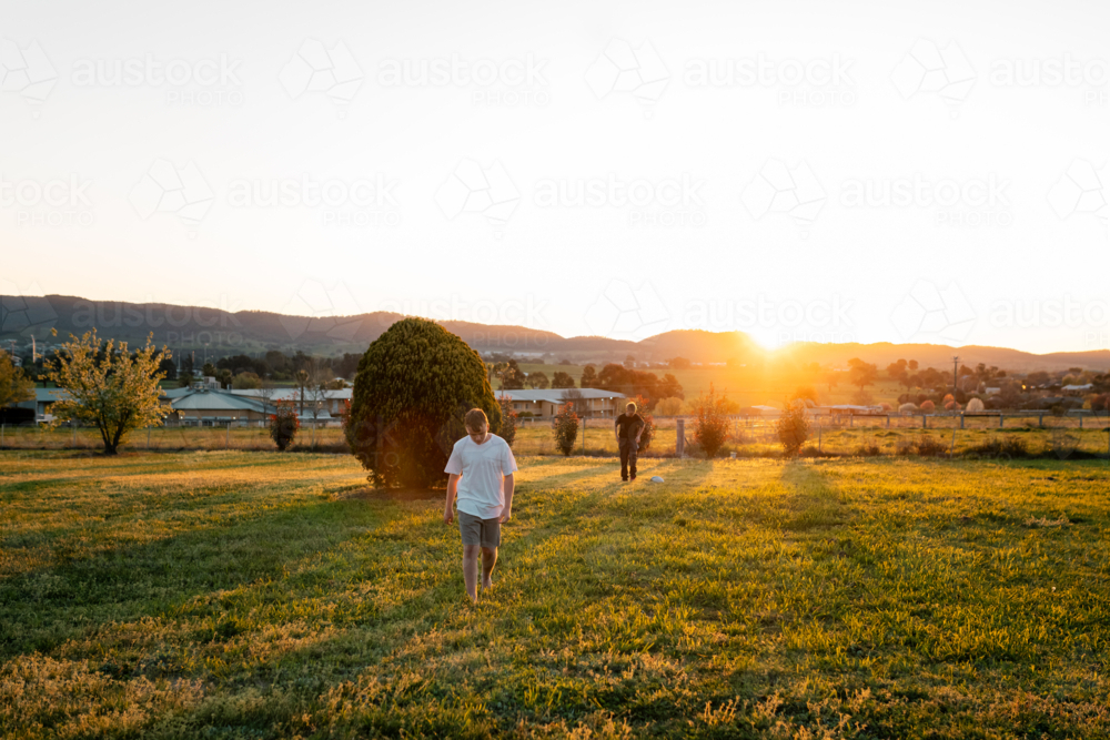 Teens walking in sunset landscape, long shadows on field, mountains in background - Australian Stock Image