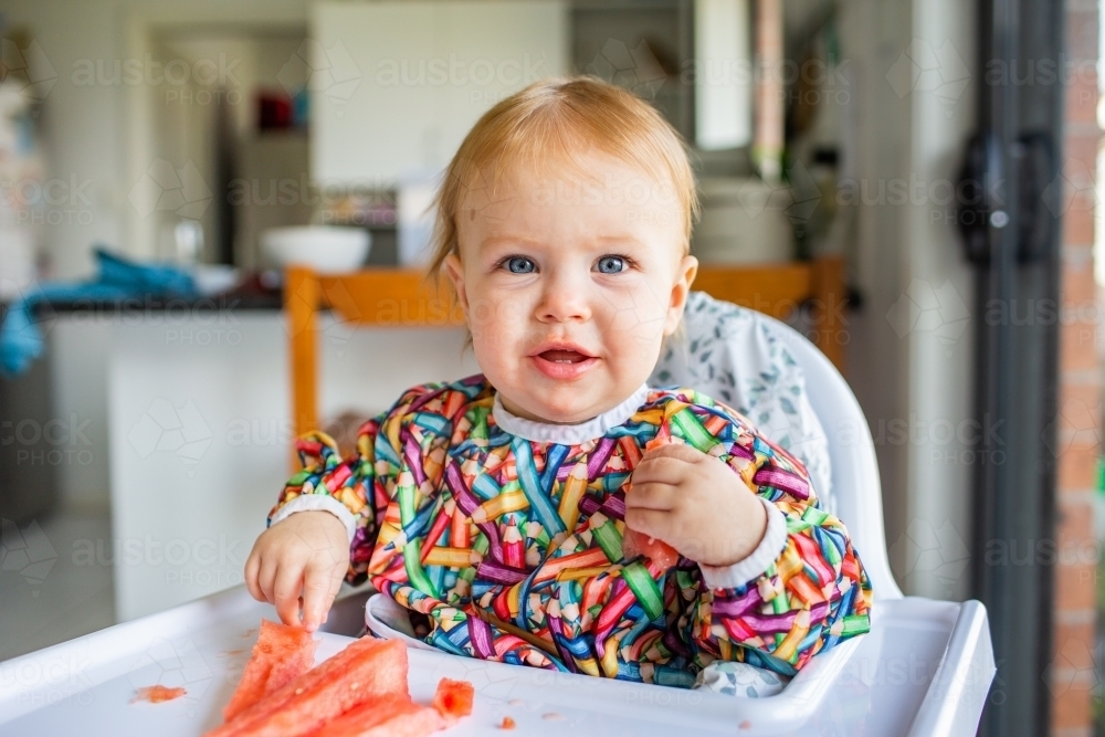 Ten month old baby eating watermelon on summer day in high chair - Australian Stock Image