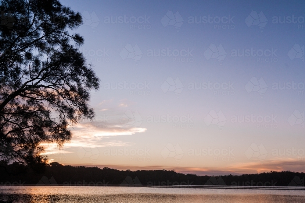 The sun sets behind the silhouette of a tree on the left overlooking a large, smooth lake. - Australian Stock Image