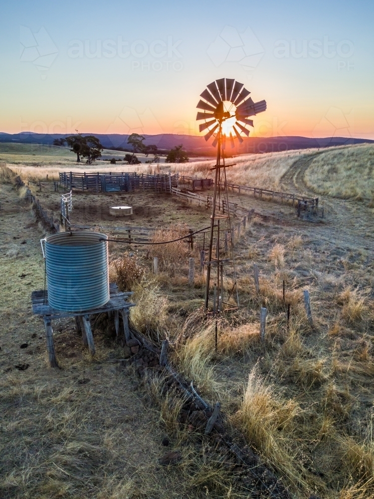 The sun setting behind a still windmill and empty tank on a dry farmland - Australian Stock Image