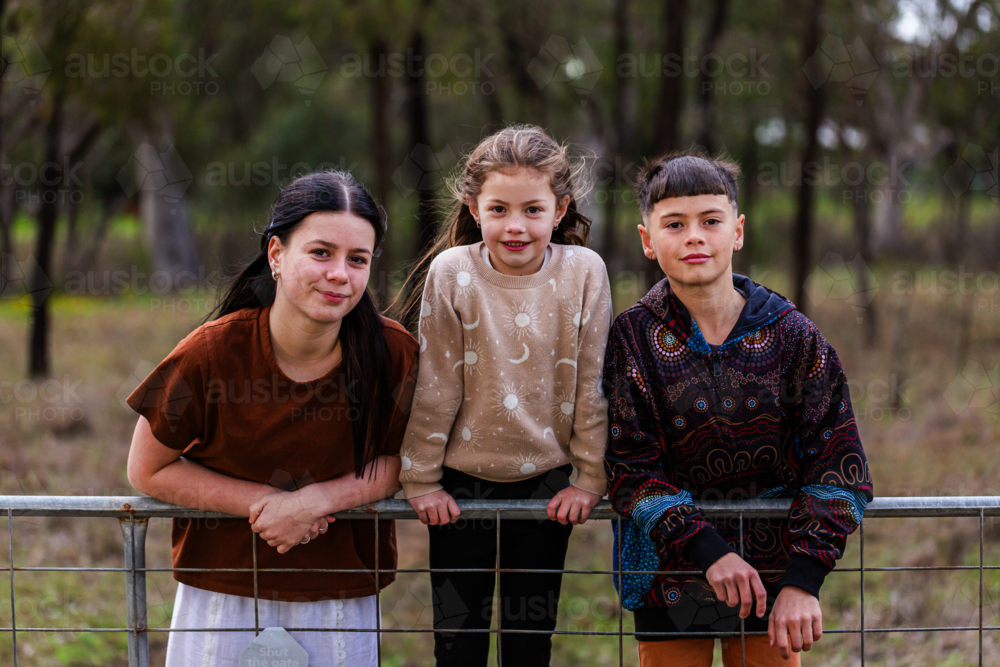 Three aboriginal kids leaning on fence together in paddock in winter - Australian Stock Image