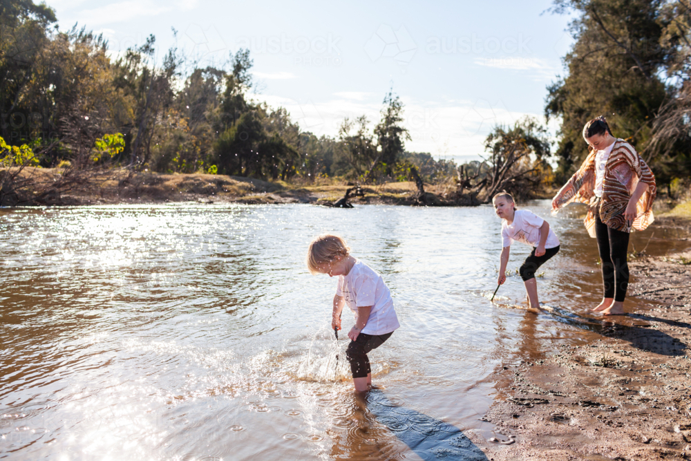 Three Aboriginal siblings playing by Hunter River in sparkling morning light toddler splashing - Australian Stock Image