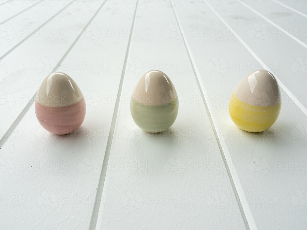 Three ceramic easter eggs with pastel stripes on white timber table top - Australian Stock Image