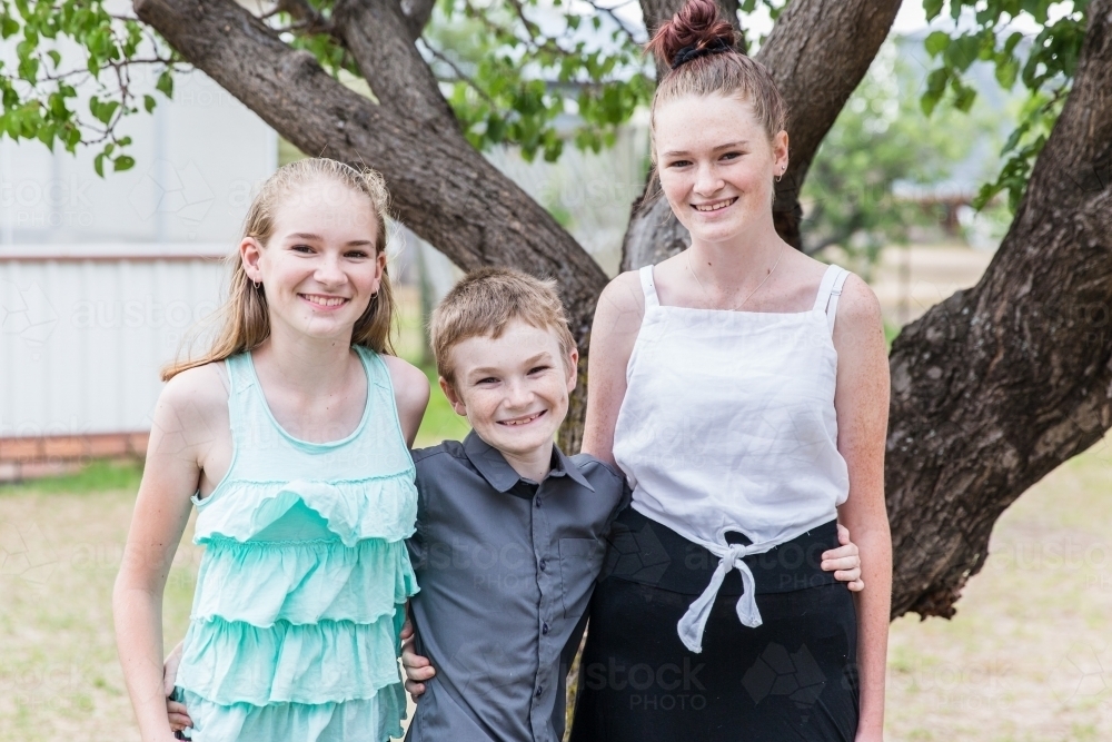 Three children brother and sister standing arm in arm in yard at home smiling happy - Australian Stock Image